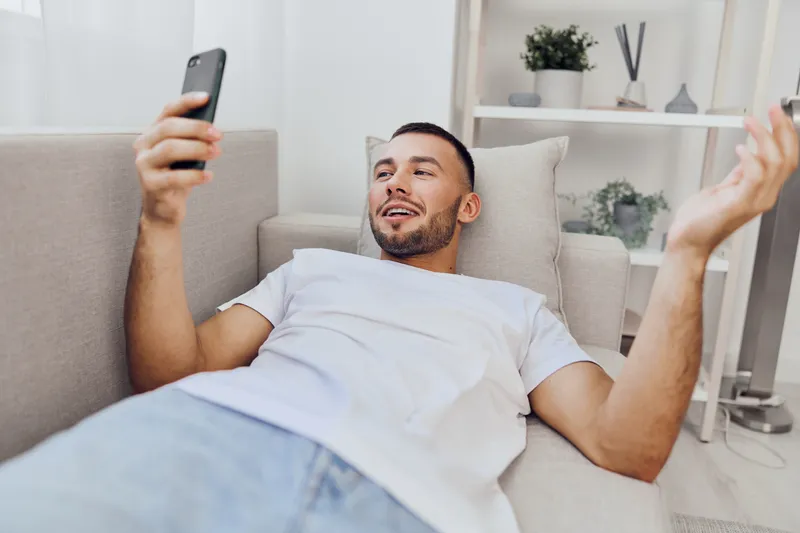 Relaxed man using smartphone at home, enjoying leisure time with natural light. Casual indoor setting with minimalistic decor for contemporary lifestyle.