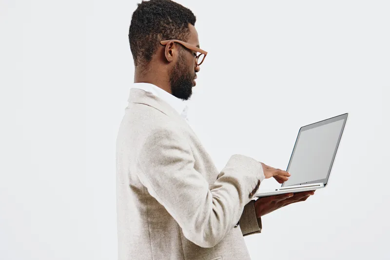 Professional young man working with a laptop, dressed in beige sweater and glasses, standing and looking at the screen, isolated on plain white background, studio shot, business and technology concept