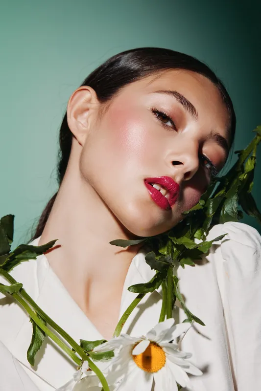 Portrait of a young woman with glossy red lips and glowing skin posing with white daisies on a green background. Beauty creative fashion concept with natural elements.