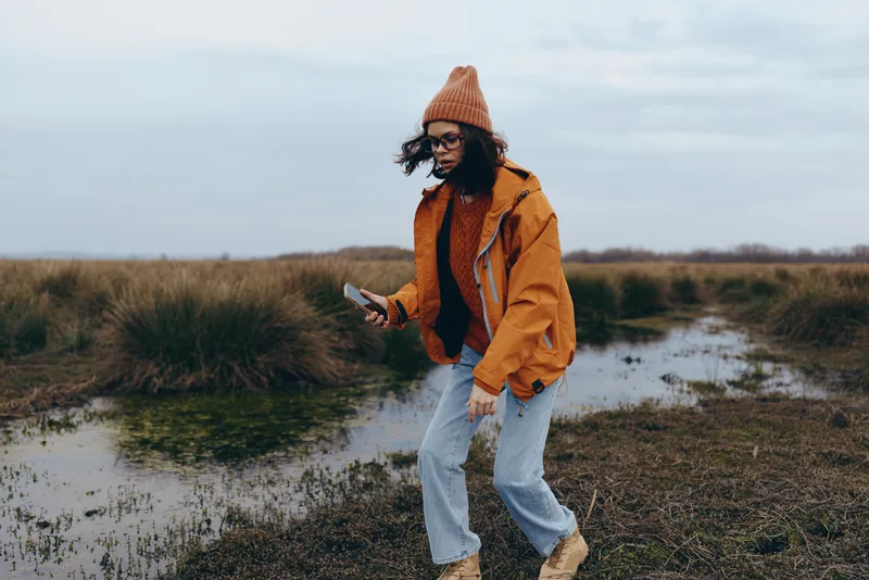 Woman in orange jacket and knit hat walks outdoors along a small river in a natural field, using a smartphone, casual lifestyle photo in cloudy countryside in autumn, alone, calm mood, soft daylight