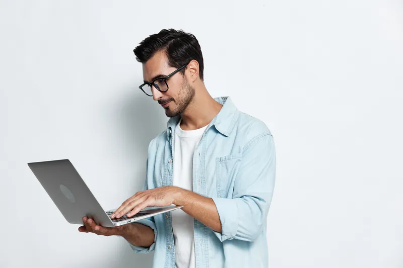 Handsome young caucasian man wearing glasses and a blue denim jacket, working on a silver laptop, looking focused and engaged against a light background. Lifestyle concept.