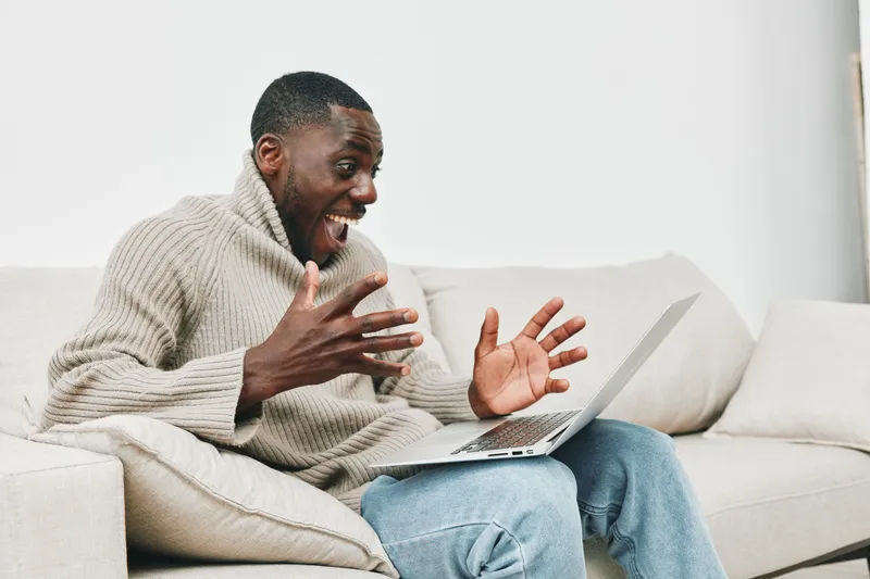 Excited African man using a laptop at home in a cozy sweater, experiencing joy while engaging online, bright atmosphere with modern decor