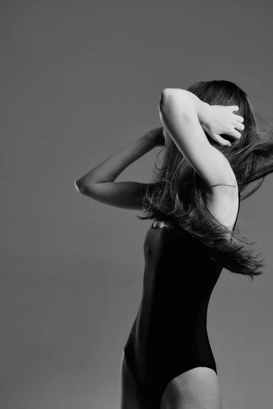 Black and white silhouette of a woman with long hair in a bodysuit, expressing motion and elegance in a minimalist studio setting.
