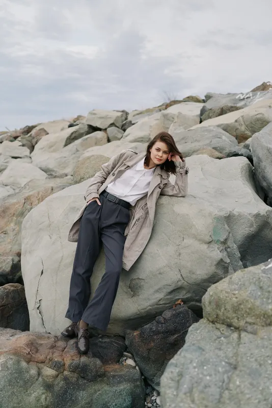 Woman in stylish coat and white shirt leans on large rocks at cloudy beach. Casual fashion outfit and natural setting create mood of relaxed outdoor lifestyle and autumn season.