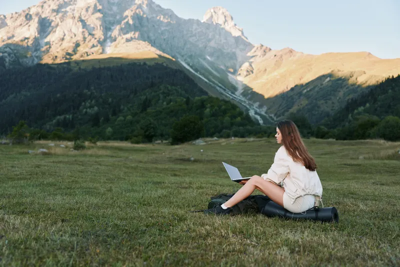 A young woman sits on a grassy field with a laptop on her lap, mountains rising in the distance under a clear sky. She focuses on the screen, portraying remote work in a natural setting.