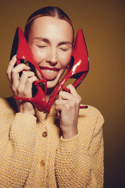 Young woman with closed eyes holds red high heels near face and sticks out tongue. Fashion concept with playful mood and bright studio lighting on yellow background.