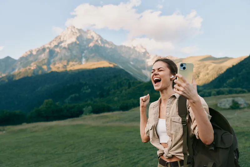 Young woman with backpack takes a joyful selfie outdoors with mountains in the background during a sunny day, expressing excitement and freedom in nature.