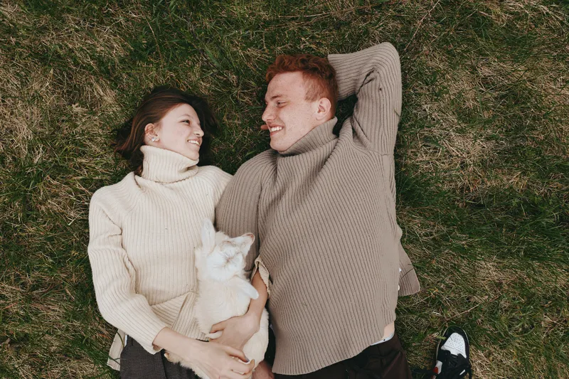 A couple lies on grass in cozy sweaters, smiling at a playful goat kid between them, capturing a warm lifestyle moment outdoors and showcasing affection, relaxation, and family connection.