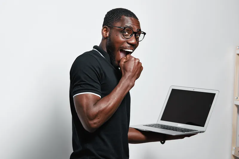 African American man with dark skin and glasses holding laptop and biting fist in excitement or surprise in studio with white background. Casual clothing and technology concept.