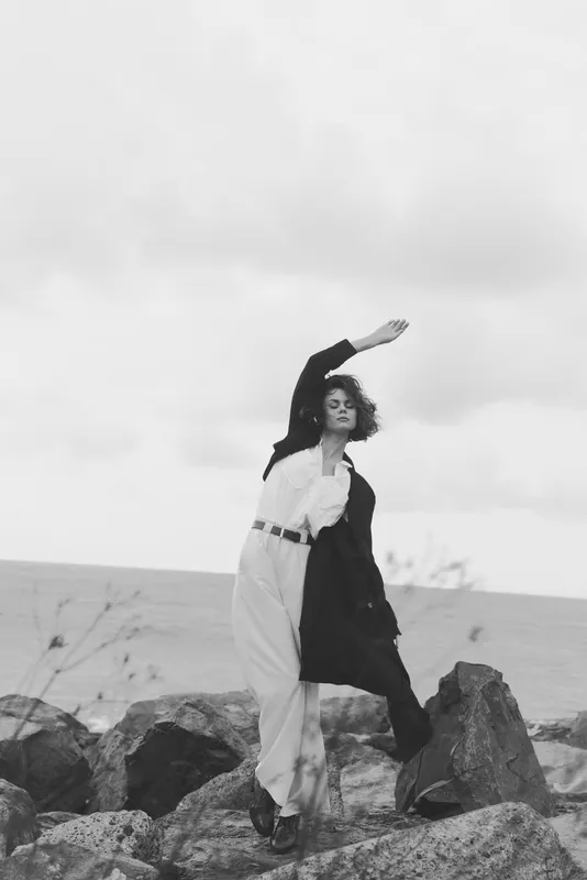 Dancer pose on a rocky shoreline with rocks, wind sweeping fabric in a dramatic monochrome outdoor shot with bold movement and artistic flair
