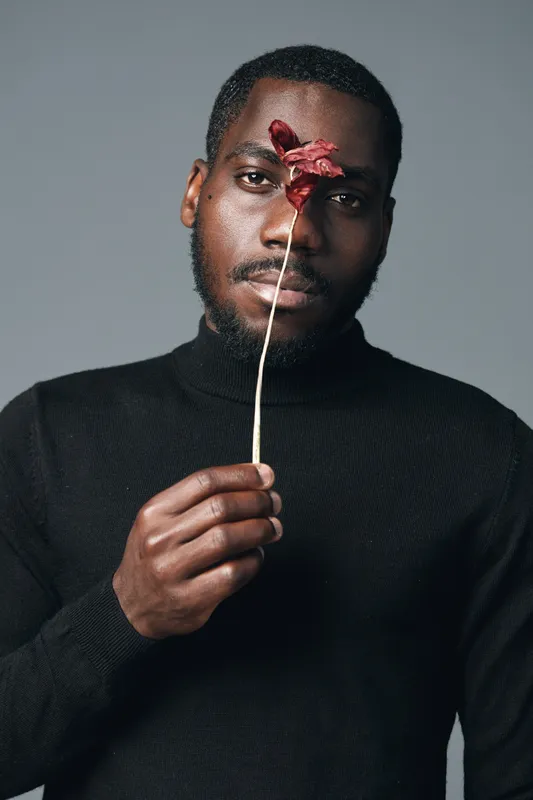 African man with dark skin holding dried flower in front of face in creative conceptual visual style. Portrait with black turtleneck and neutral background for artistic expression and emotion.