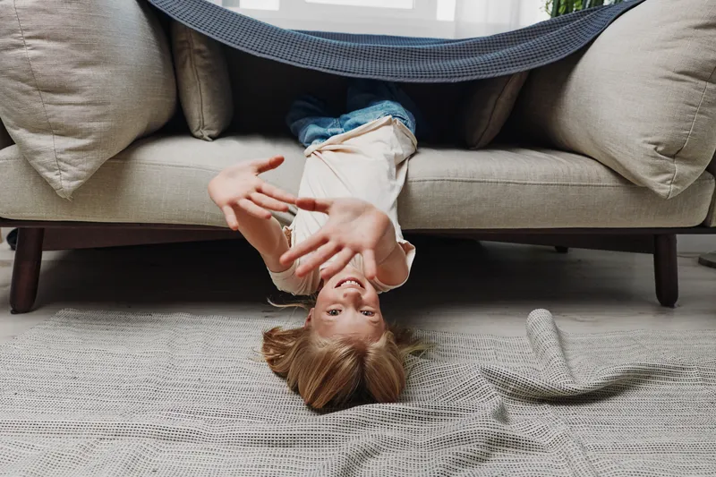 Happy child plays upside down under couch with blanket hammock in cozy living room, smiling and reaching hands forward. Concept of childhood fun and imagination.