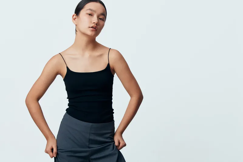 Young woman in black tank top and gray pants stands confidently with hands in pockets on white background, posing for fashion portrait and casual style studio shoot.