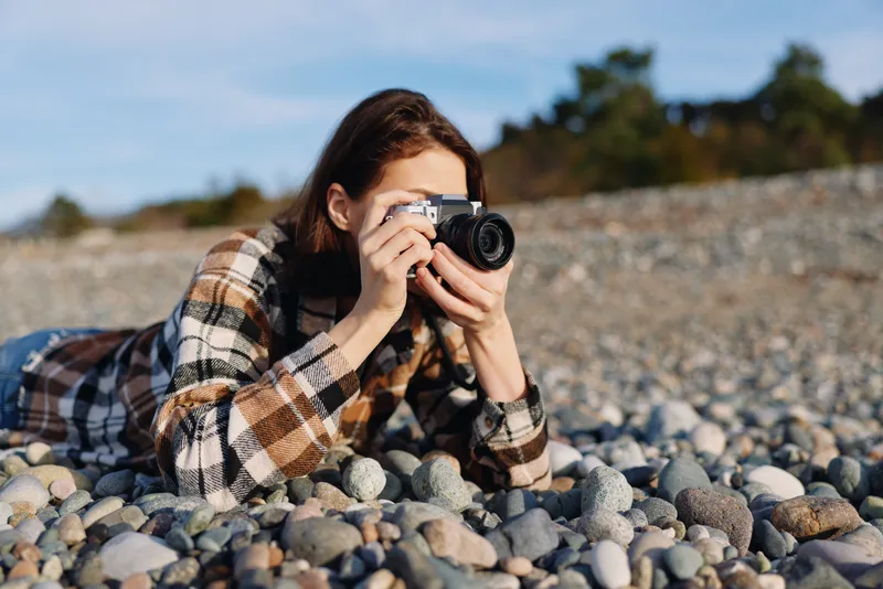 Photographer lying on a rocky beach in a plaid shirt, aiming a camera toward the horizon for a candid outdoor landscape shot with natural light and coastal texture.