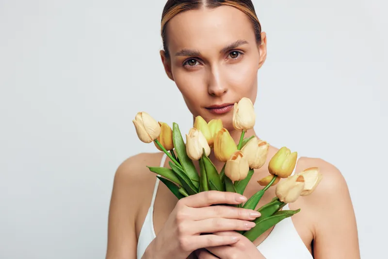 Young woman holding bouquet of yellow tulips close to her face, soft natural light, neutral background, calm expression, minimalistic style, fresh spring mood