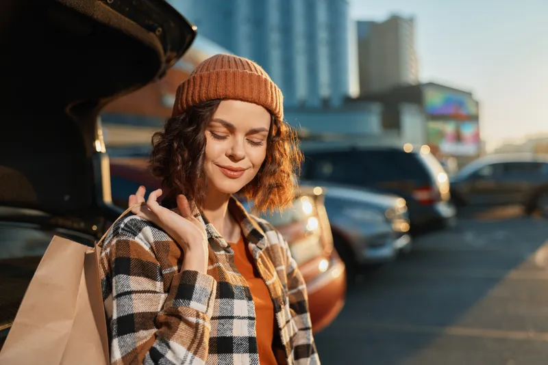 Woman shopping by car in urban parking lot, holding bags and smiling in golden hour glow; candid portrait with authenticity, mindful living and emotional storytelling vibe.