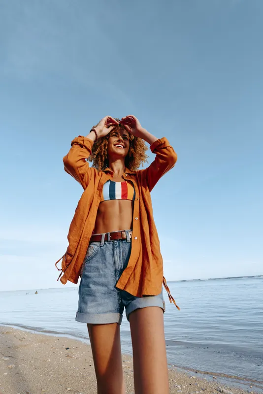 Young woman with curly hair enjoys a sunny day at the beach wearing a striped bikini top and an orange shirt over denim shorts with a joyful smile.