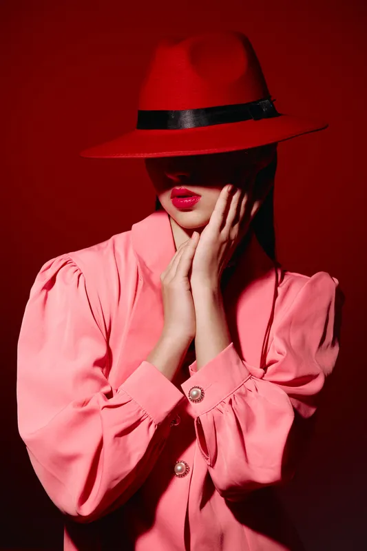 Stylish woman in a red fedora hat covers eyes partially with pink blouse and red lipstick. Studio shot on intense red background. Creative beauty and fashion concept.