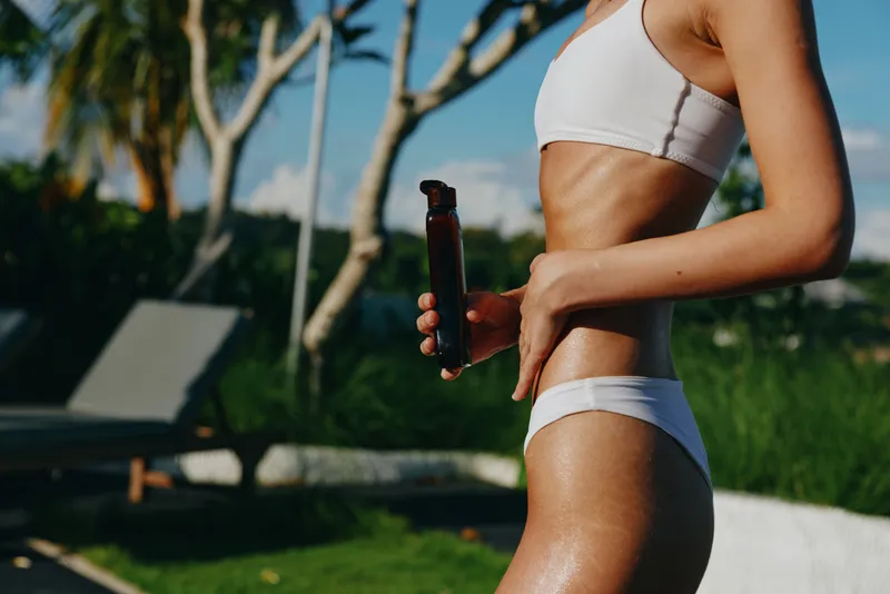 Cheerful woman outdoors in a bikini, holding a bottle by a pool, sunlit skin and relaxed posture, outdoor lifestyle moment with bright greenery, summer sunshine and candid wellness vibe