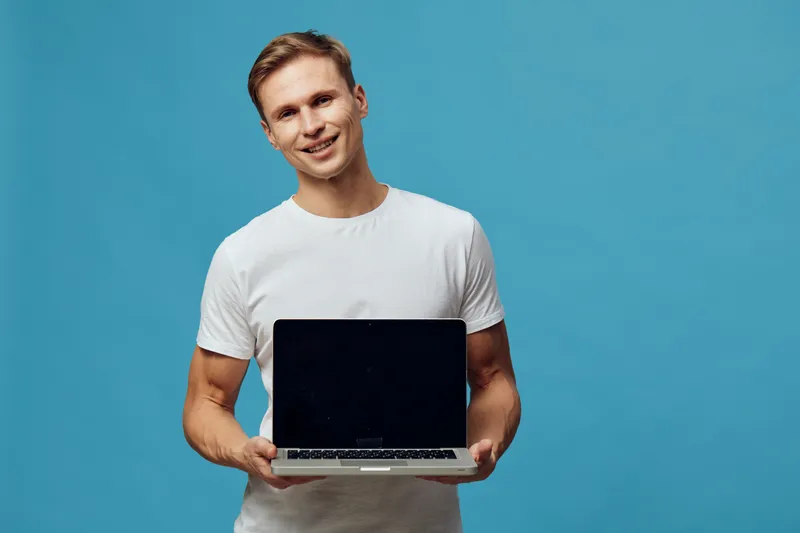 Smiling young man in casual white t-shirt holding open laptop computer with blank black screen, looking at camera with friendly expression, isolated on solid blue background, studio portrait. People l
