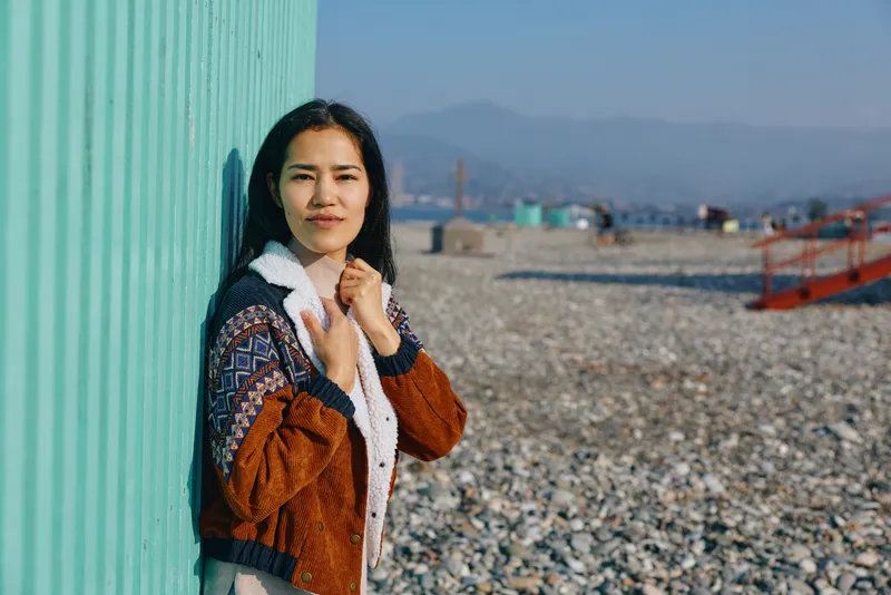 Beachside scene with a woman standing beside a green wall, wearing a brown jacket and white fleece, pebbled shore and distant hills in the background, casual outdoor fashion