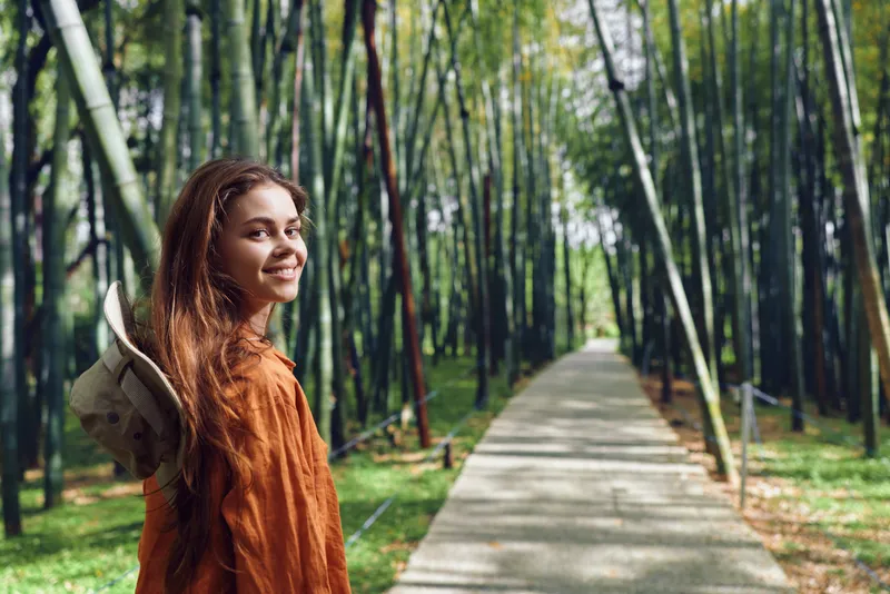 Woman walking on a paved path through bamboo forest, portrait of traveler with backpack and hat, smiling and looking back with a warm smile as sunlight lights the green outdoor scene