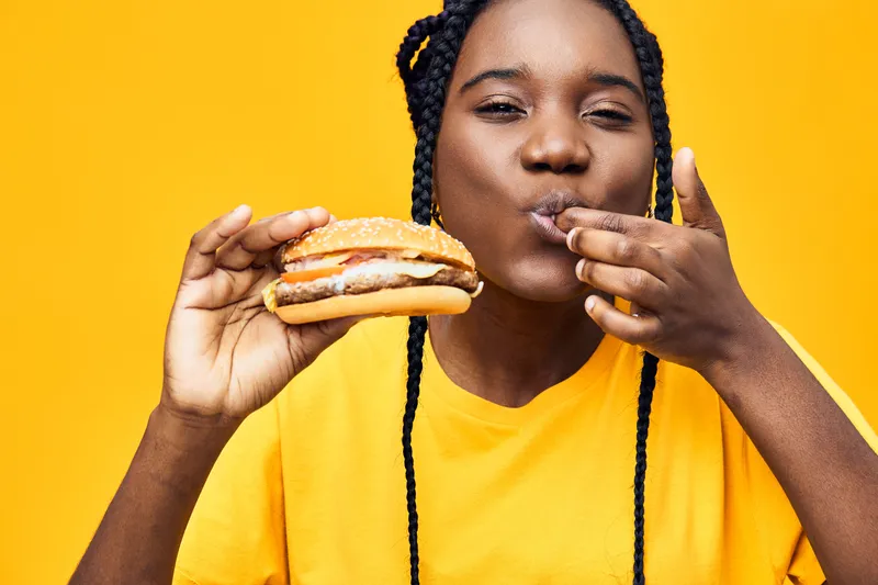Happy young woman enjoying a delicious burger against a vibrant yellow background, showcasing her love for fast food and cheerful emotions