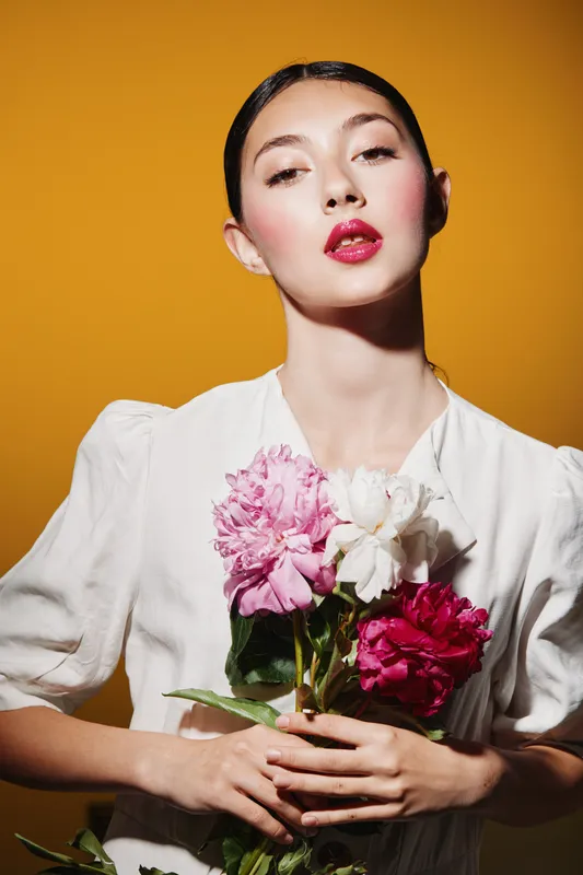 Young stylish woman with vibrant makeup and pink lipstick wearing a white blouse holds pink and white peonies against a warm yellow background. Beauty fashion creative portrait.