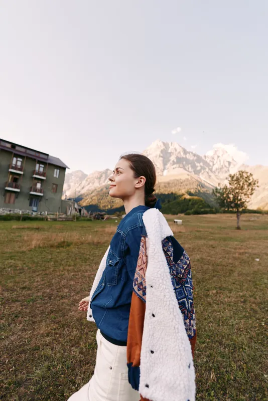 Woman walking in field with jacket draped over shoulders, profile portrait outdoors near mountains. Casual denim shirt, relaxed smile, autumn landscape and alpine scenery.