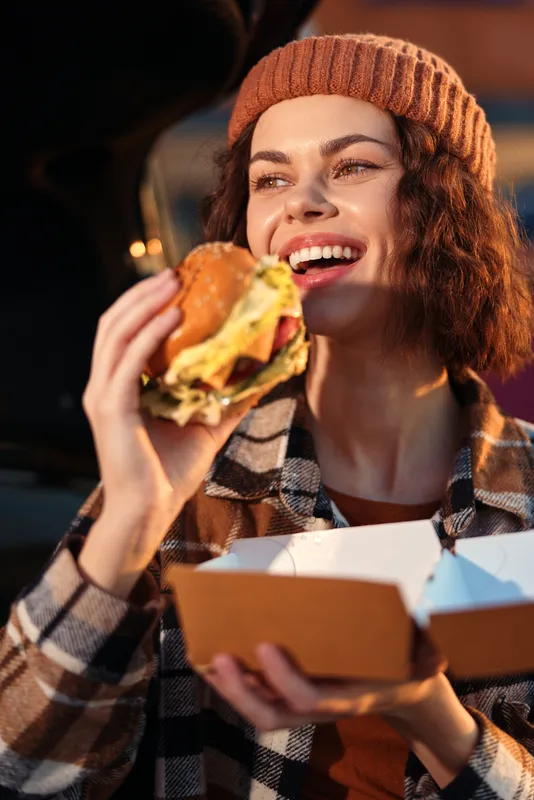 Woman smiling as she enjoys a burger and street food outdoors, urban evening takeout moment with beanie and plaid jacket. Candid lifestyle shot with authenticity, golden hour glow and emotional