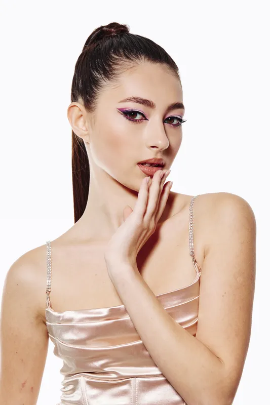 Young woman in shiny satin dress poses with elegant hand near face. She wears creative soft makeup with bright eyeshadow. Studio shot on white background.