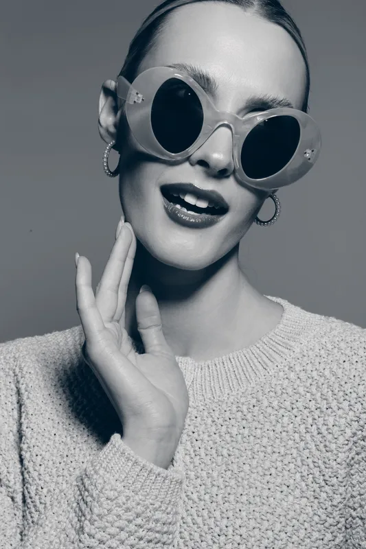 Young caucasian woman with fair skin wears large round sunglasses and earrings. She smiles with slightly open mouth and touches her face. Concept of fashion, style, and beauty.