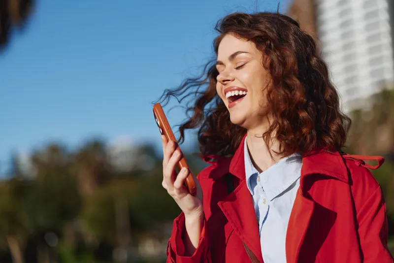 Young woman with curly hair smiles while checking her phone outdoors, wearing a red jacket under a bright blue sky.