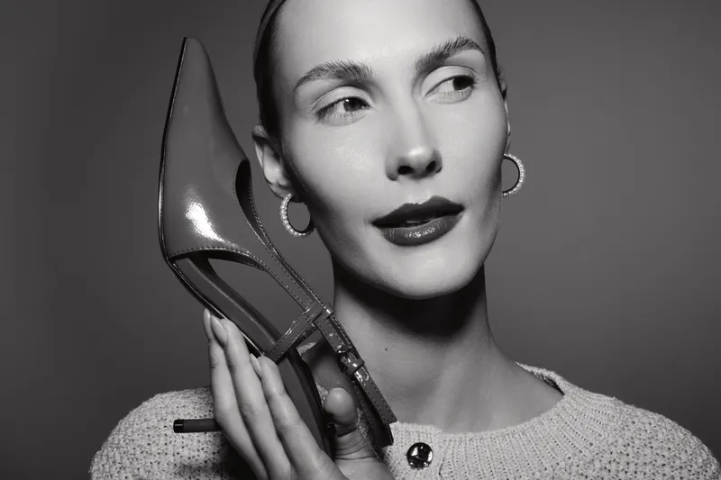 Portrait of a fair skinned caucasian woman holding a high heel shoe near her face, wearing hoop earrings and a textured sweater in a studio setting.