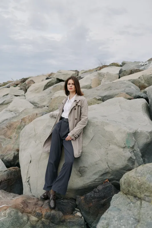 Young woman with long brown hair wearing a beige coat and dark trousers stands on rocky beach under cloudy sky. Casual fashion portrait with natural background and moody atmosphere.