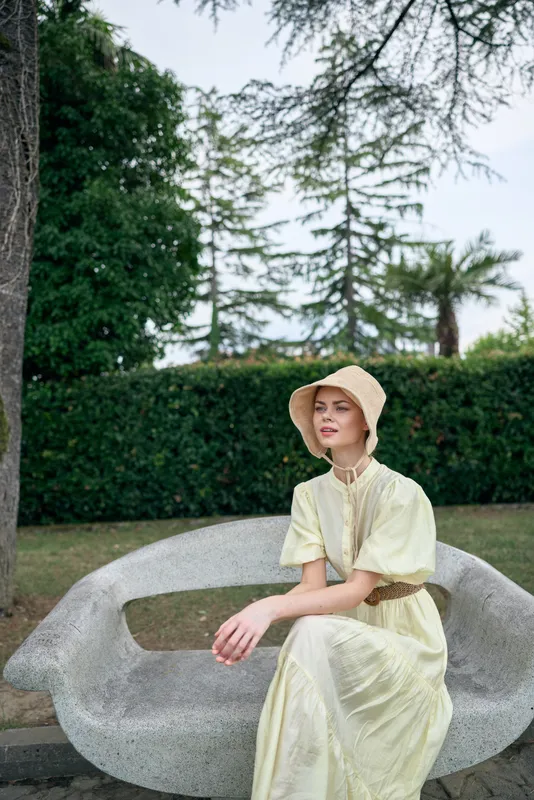 woman wearing a pale dress and hat sits on a sculptural bench in a park, serene outdoor setting, showcasing fashion and calm demeanor in natural light.