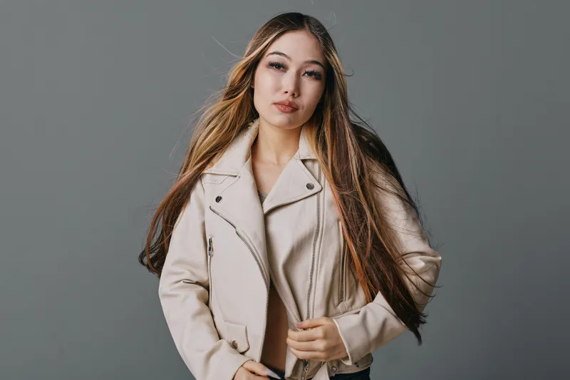 Young woman with long hair wearing beige leather jacket posing against gray background, looking confident and stylish with natural makeup and casual outfit in studio portrait.
