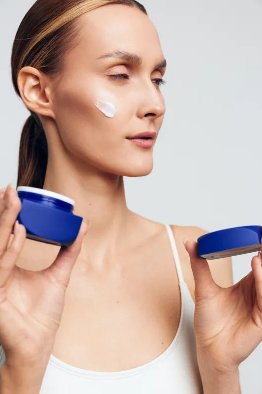 Young woman with clear skin applying white face cream on cheek, holding blue jar. Concept of skincare, beauty, self-care, and moisturizing with soft natural lighting on white background.
