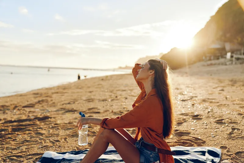 Woman in an orange top sits on a towel at the beach, holding a water bottle and gazing at the sun. The scene presents outdoors, a relaxing vibe and summery scenery.