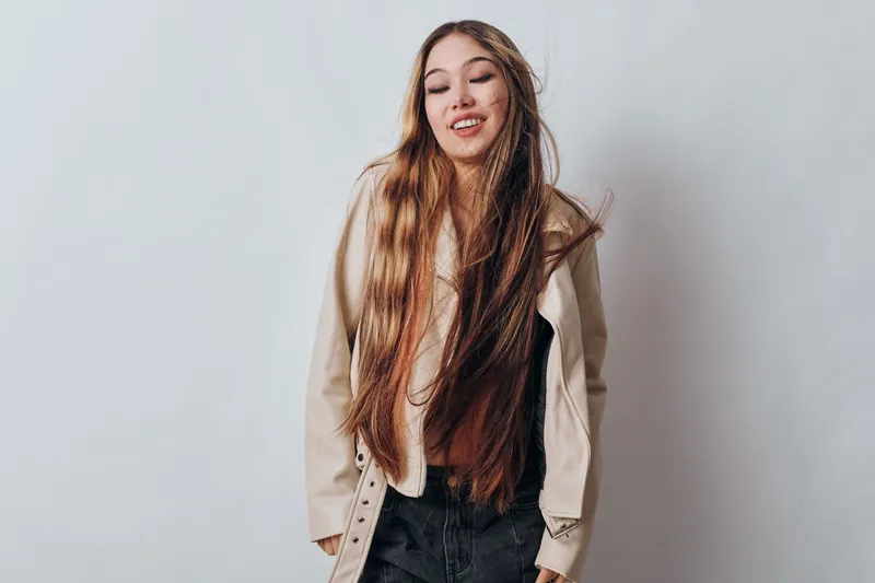 Young woman with long hair wearing casual outfit and light jacket standing against white background with eyes closed and relaxed expression. Female, fashion, portrait, model.