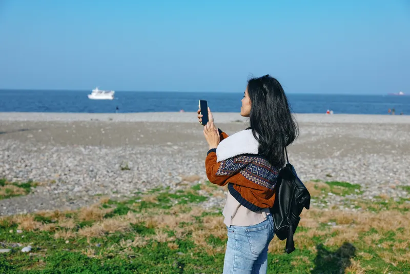 Woman on a sunny beach holding a smartphone to capture the seascape, backpack on her shoulder, casual clothing, salty breeze, wide horizon, relaxed travel moment, outdoor photography by the shore