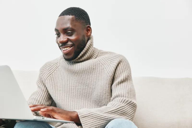 Happy young Black man in a cozy sweater using a laptop, expressing joy and engagement Perfect for concepts of remote work and digital communication