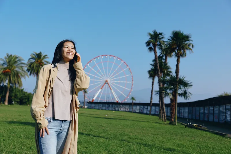 Woman stands outdoors in a park beside a ferris wheel, wearing casual fashion and a light coat, smiling while talking on her phone under sunny skies amid tall palm trees.