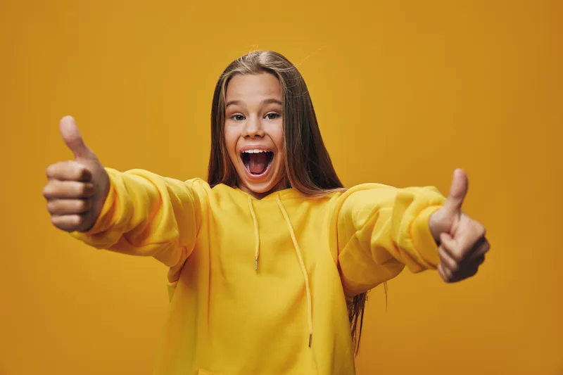 Girl in yellow hoodie showing both thumbs up with excited open mouth expression on yellow background for positivity and success feeling.