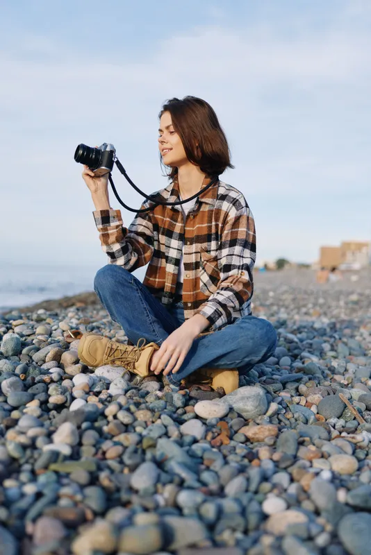 Photographer on a pebble beach, outdoor casual style, sits with a camera by the sea, enjoying sunlight and breeze while capturing textures and gentle waves in a peaceful seaside moment