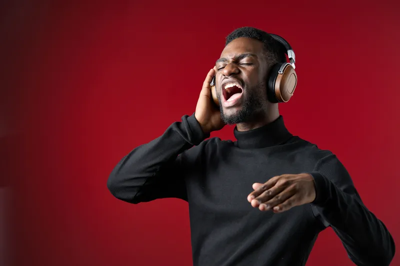 Young Black man passionately singing with headphones against a vibrant red background, portraying joy and music enjoyment, ideal for music and lifestyle themes