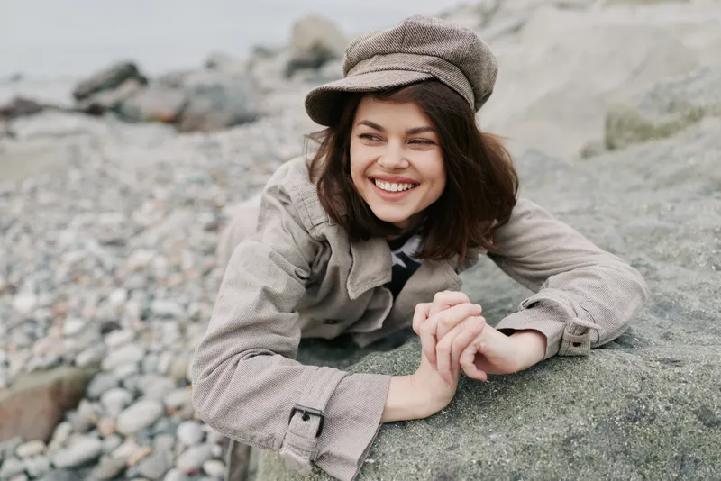 Smiling young woman in a coat and hat leans on a rock at a rocky beach, enjoying a relaxed moment outdoors. Natural light highlights her joyful expression and casual style.