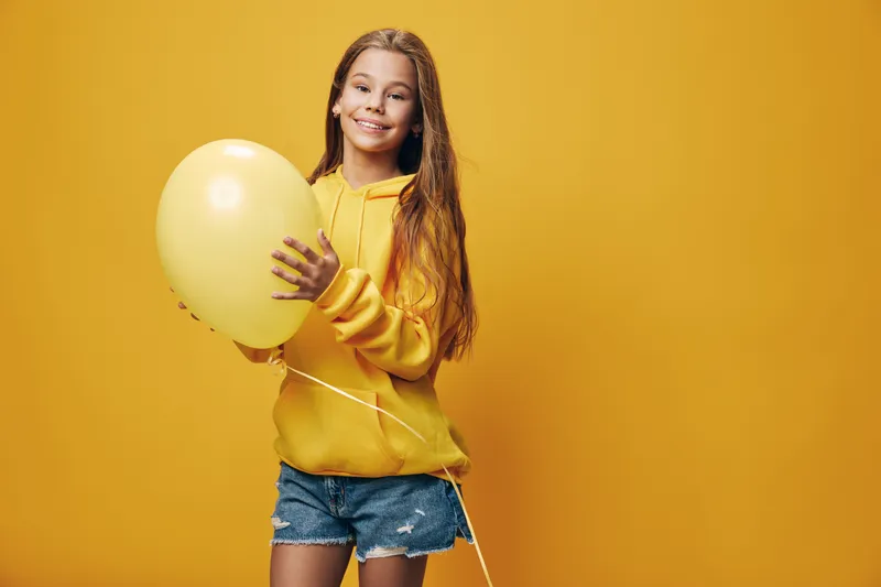 Girl wearing yellow hoodie and denim shorts smiling with yellow balloon standing against yellow background in studio. Child holding balloon with happy expression for celebration.