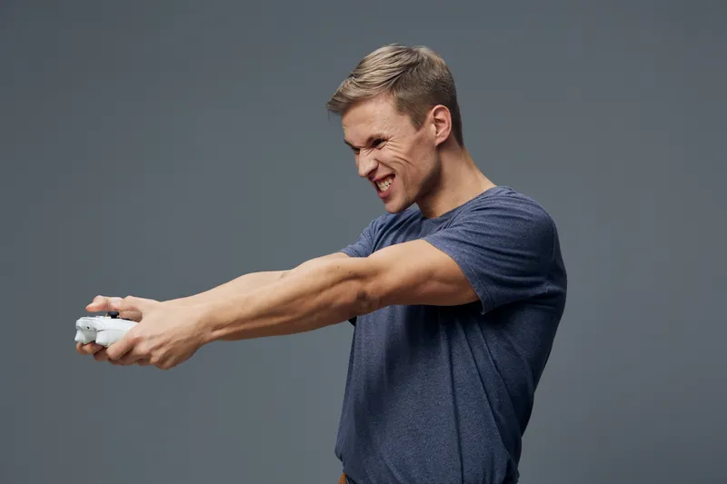 Active young Caucasian man with short hair and a broad smile, wearing a casual blue T-shirt, stretching his arms forward holding a gaming controller, isolated on a plain gray background, studio shot. 
