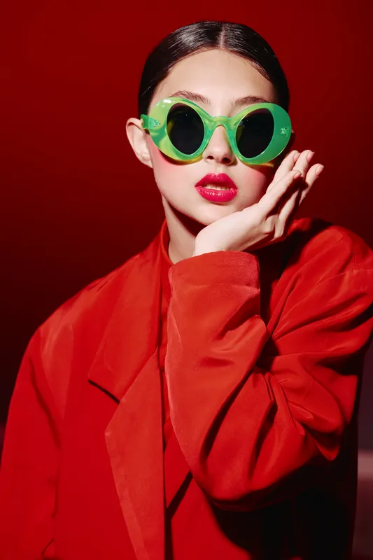 Fashionable woman with slicked dark hair wears vivid green glasses and bright red lipstick, dressed in oversized red clothing against a matching red background. Trendy beauty concept.